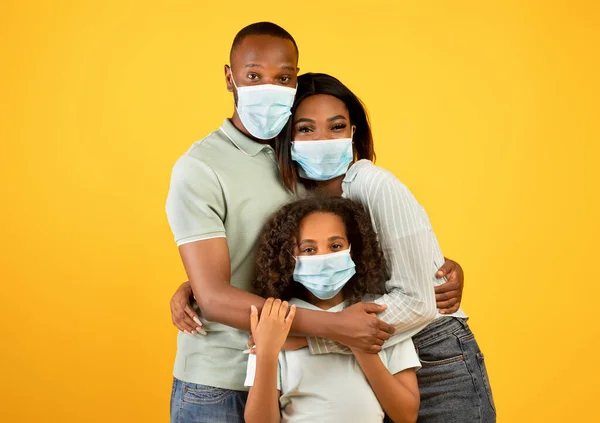 Jóvenes padres afroamericanos y su hija con máscaras médicas protectoras, abrazando sobre fondo amarillo —  Fotos de Stock