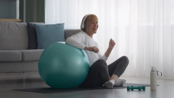 Mujer mayor escuchando música usando auriculares cerca de Fitball en casa — Vídeos de Stock