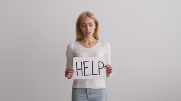 Жертва домашнього насильства. Молода нещасна жінка сумно позує на білому тлі, піднімаючи плакат зі словом HELP — стокове відео