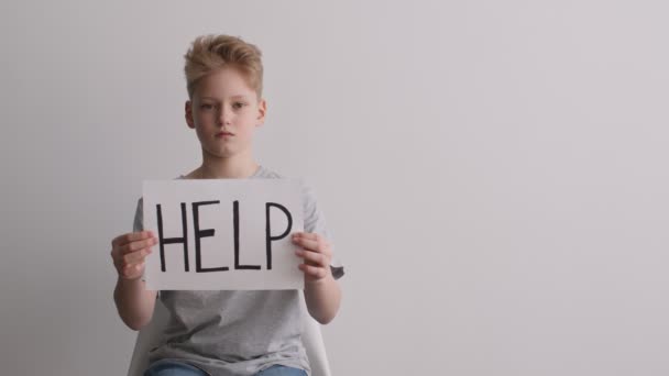 Roep om hulp. Studio portret van ongelukkige tiener jongen tonen woord HEPL op poster, witte muur achtergrond, lege ruimte — Stockvideo