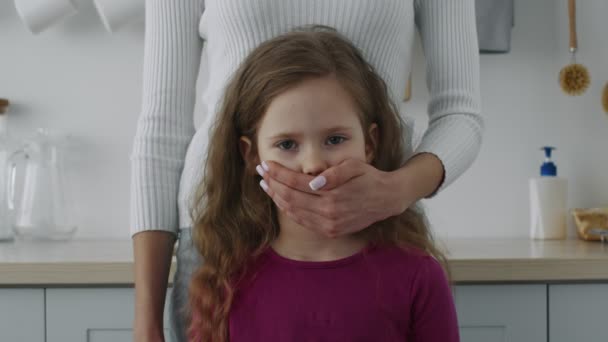 Секреты семейной жизни. Портрет испуганной маленькой девочки, смотрящей в камеру, руки матерей, закрывающие рот — стоковое видео