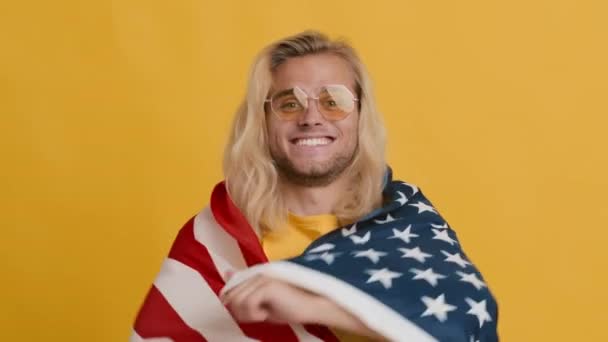 Красивый длинноволосый парень, прикрывающий себя флагом США и улыбающийся в камеру — стоковое видео