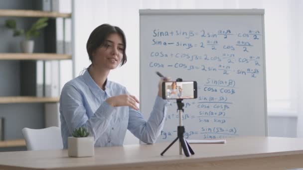 Jeune professeur de mathématiques joyeux enregistrement vidéo pour blog sur smartphone, assis à la salle de classe avec des formules mathématiques — Video