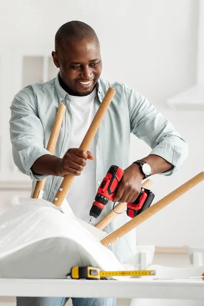 Χειροκίνητη εργασία στο σπίτι. Χαρούμενη χιλιετή μαύρο άνδρα συναρμολόγηση επίπλων με ηλεκτρικό κατσαβίδι στο εσωτερικό της κουζίνας, καλλιέργεια — Φωτογραφία Αρχείου