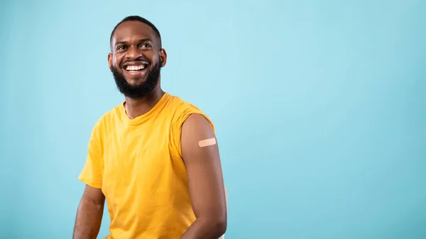 Hombre negro vacunado alegre con vendaje de yeso en el hombro después de la inyección de la vacuna covid-19 sobre fondo azul —  Fotos de Stock