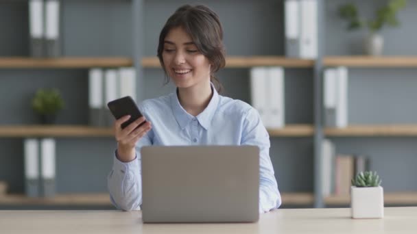 Güzel telefon. Ofiste dizüstü bilgisayarda çalışan, cep telefonuna bakan ve telefona cevap veren genç pozitif bir kadın. — Stok video