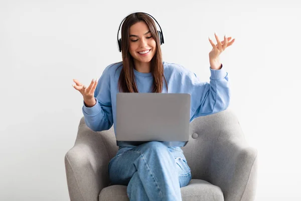 Mujer joven feliz en los auriculares que tienen reunión de negocios en línea en el ordenador portátil, sentado en cómodo sillón sobre fondo blanco — Foto de Stock
