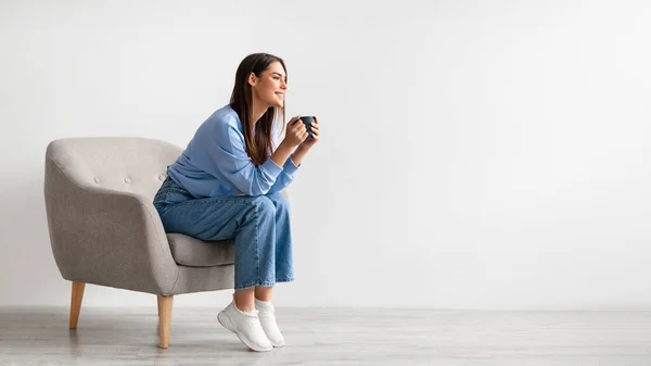 Longitud completa de la mujer joven que tiene descanso de café en acogedor sillón contra la pared del estudio blanco, pancarta con espacio libre —  Fotos de Stock