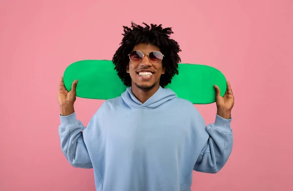 Adolescente preto encaracolado positivo com deck de skate sorrindo para a câmera no fundo do estúdio rosa. Equipamento desportivo — Fotografia de Stock