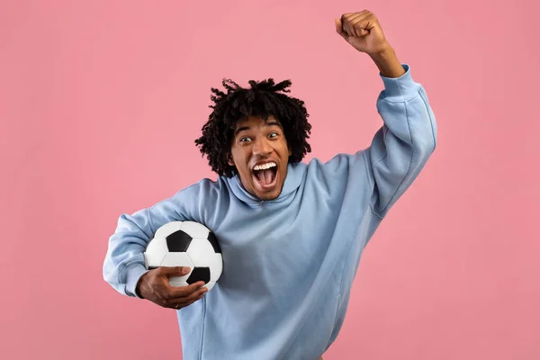 Feliz adolescente negro com bola de futebol torcendo por seu time de futebol favorito no fundo do estúdio rosa — Fotografia de Stock