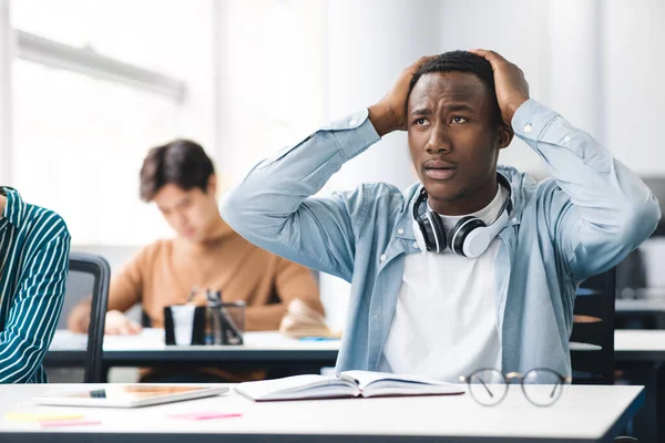 Черный студент хватает голову, сидя за столом в классе — стоковое фото