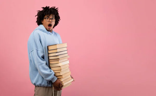 Nerdy preto adolescente estudante sentindo-se chocado com pilha de lição de casa, segurando grande pilha de livros em fundo rosa — Fotografia de Stock