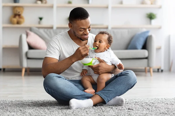 Cuidar a papá negro alimentando a su adorable bebé desde la cuchara en casa —  Fotos de Stock