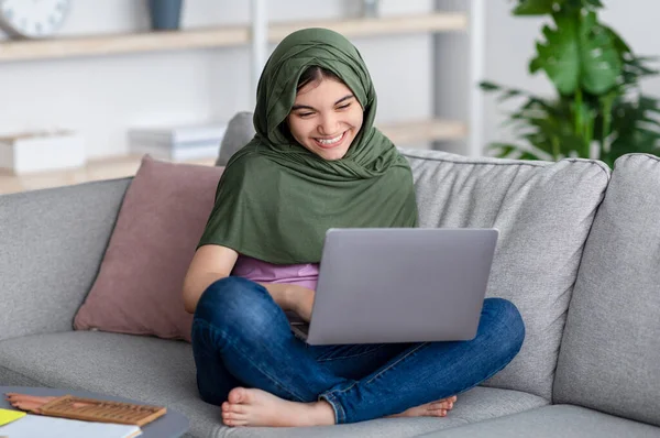 Concepto de aprendizaje remoto. Feliz chica adolescente musulmana en hijab estudiar en línea, sentado en el sofá con el ordenador portátil en casa — Foto de Stock