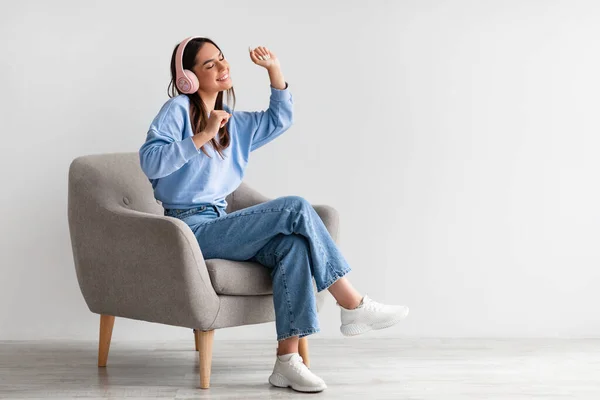 Feliz dama milenaria en auriculares inalámbricos relajándose en sillón y disfrutando de la música contra la pared blanca del estudio — Foto de Stock