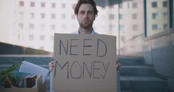 Zoom in portrait of young workless manager sitting outdoor with poster Need Money, ψάχνει για νέα δουλειά, αργή κίνηση — Αρχείο Βίντεο