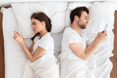 Evde sırt sırta yatarak cep telefonu kullanan çift.