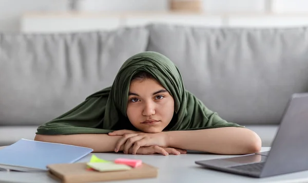 Muchacha adolescente musulmana en hijab acostado en la mesa con el ordenador portátil y materiales de estudio, tener lección remota aburrida, aprender en línea — Foto de Stock