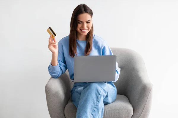 Kredi kartı ve dizüstü bilgisayarı olan genç bir kadın rahat koltukta oturuyor, internetten mal sipariş ediyor, web mağazasından bir şeyler alıyor. — Stok fotoğraf