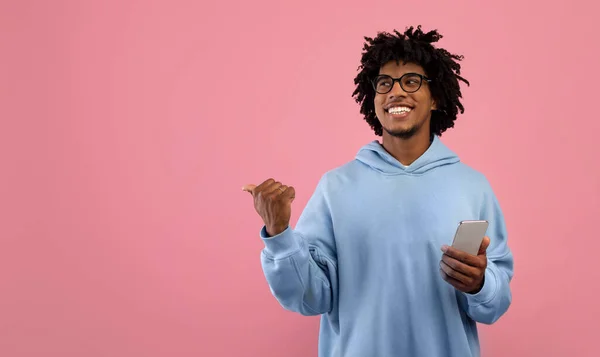 Homem negro alegre com telefone celular apontando para o espaço em branco para o seu anúncio no fundo do estúdio rosa — Fotografia de Stock