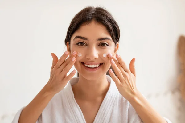 Mujer alegre aplicando crema facial hidratante de la piel posando en el baño —  Fotos de Stock