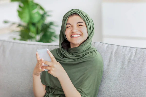 笑顔イスラム教徒の青少女でオンラインチャット,座っています上のソファとともにスマートフォン-ホーム — ストック写真