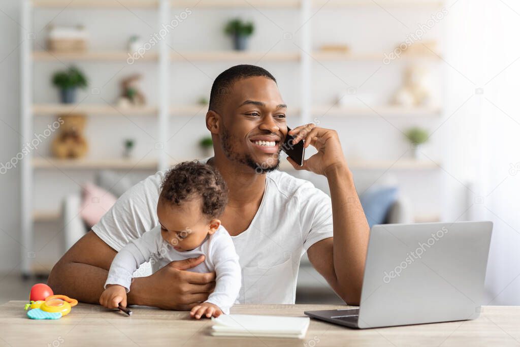 Black working father talking on cellphone while babysitting infant child at home