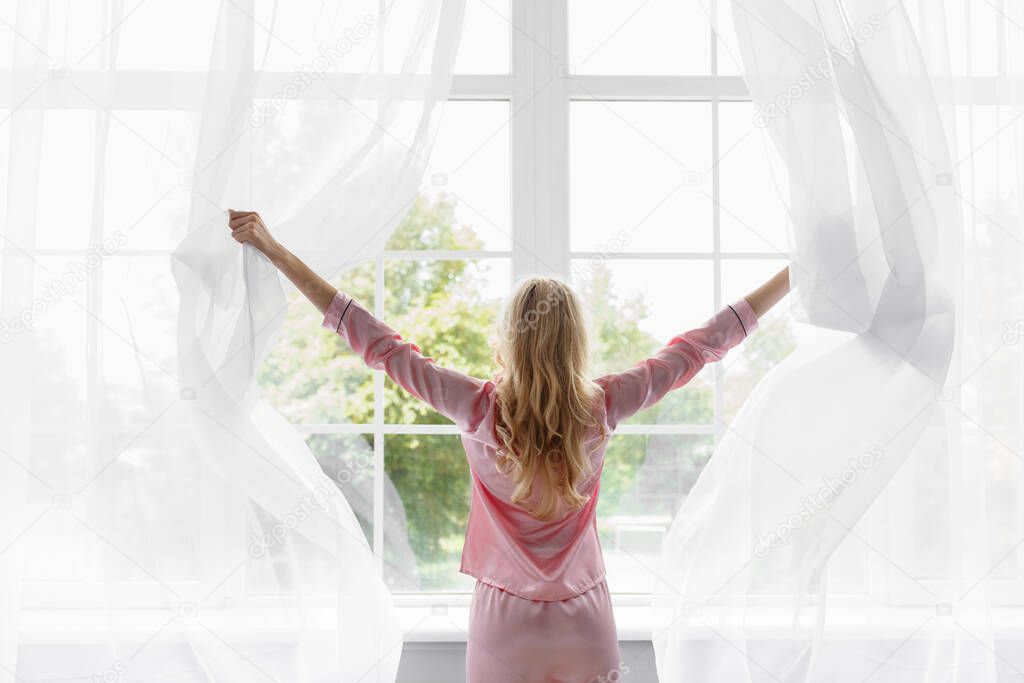 Young pretty blonde woman in pink pajamas woke up, look at large panoramic window in bedroom