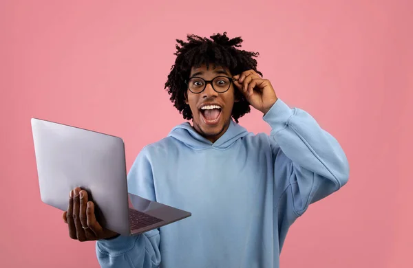 Schockierter schwarzer Teenager in lässigem Outfit blickt auf Laptop, begeistert über seinen Sieg oder Erfolg auf rosa Studiohintergrund — Stockfoto