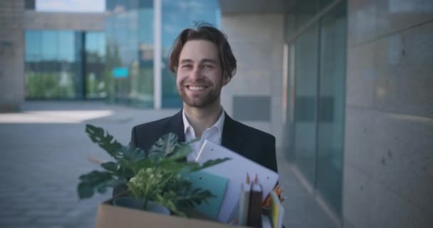 Young happy man office manager verlaten gehate baan, het dragen van doos met bezittingen, langs business center en glimlachen — Stockvideo