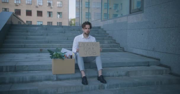 Jovem deprimido demitido gerente de escritório sentado na área de negócios com coisas pessoais e cartaz Procurando um emprego — Vídeo de Stock