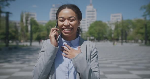 Retrato al aire libre de una joven afroamericana positiva hablando por celular, yendo a trabajar en un parque urbano — Vídeos de Stock
