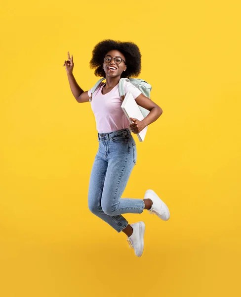Desfrutando da vida estudantil. Retrato de comprimento total da senhora americana africana pulando, usando mochila e segurando blocos de notas — Fotografia de Stock