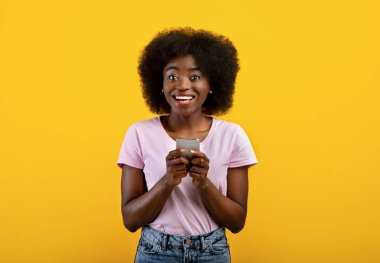 Yeni havalı uygulama. Akıllı telefonunu kullanıp gülümseyen, sarı arka planda duran mutlu zenci kadın portresi.