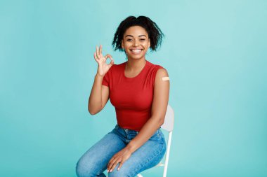 İğne yaptıktan sonra kolunda aşı olan neşeli, genç siyahi bir bayan kameraya gülümsüyor ve jest yapıyor.