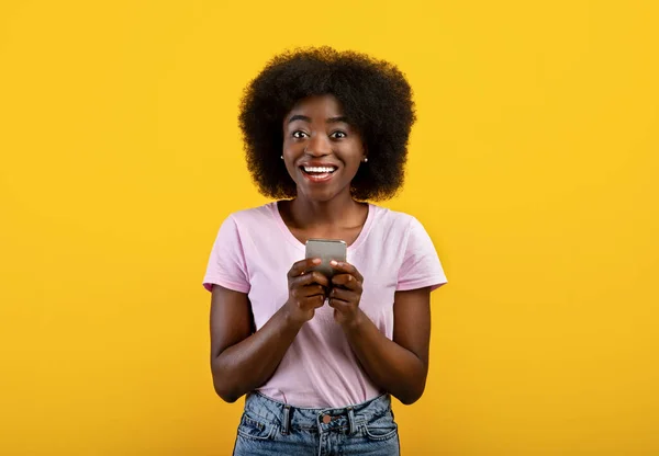 新しいクールなアプリケーション。彼女のスマートフォンと笑顔を使用して幸せな黒人女性の肖像画,黄色の背景の上に立って — ストック写真