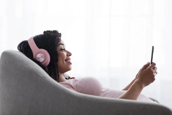 Sorridente giovane donna nera calma in cuffie wireless digitando su smartphone sul divano — Foto Stock