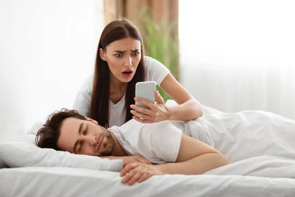 Esposa leyendo maridos mensajes en el teléfono inteligente mientras duerme en interiores — Foto de Stock
