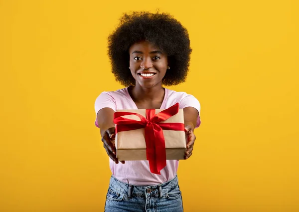 このプレゼントはあなたのためです。興奮した黒の女性の肖像画は、カメラ、黄色の背景にそれを与える包まれたギフトボックスを保持 — ストック写真
