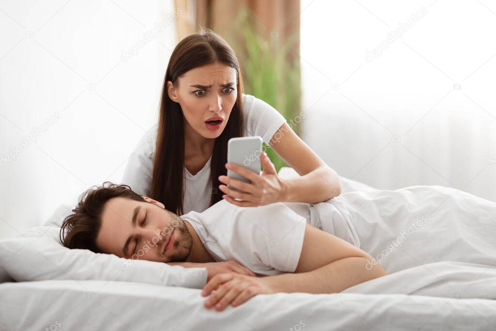 Wife Reading Husbands Messages On Smartphone While He Sleeping Indoor