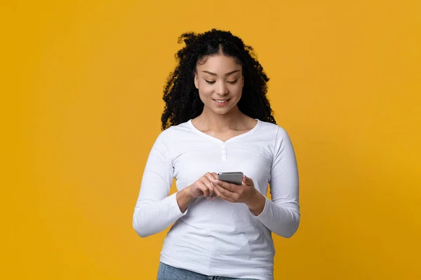 Jonge glimlachende Afro-Amerikaanse vrouw met behulp van Smartphone terwijl Staande Over Yellow Studio Achtergrond — Stockfoto