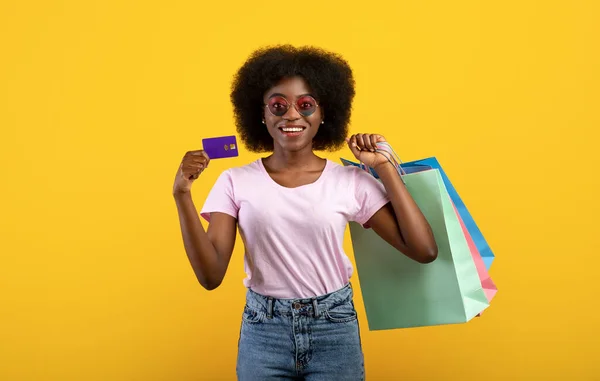 買い物が楽だ。興奮したアフリカ系アメリカ人の女性がクレジットカードと買い物袋を持って、黄色のスタジオの背景の上にポーズ — ストック写真