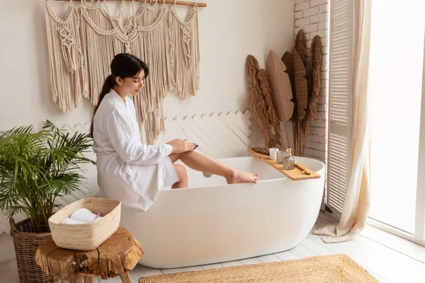 Vista lateral de la mujer que aplica la crema de depilación en las piernas en el baño —  Fotos de Stock