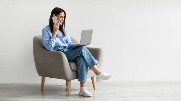 Полная длина кавказской леди с помощью ноутбука и говорить на смартфоне, сидя в кресле против белой стены — стоковое фото