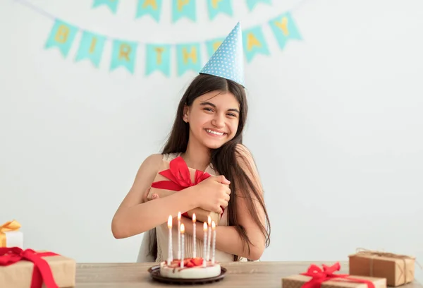 Glad indisk tonåring flicka i fest hatt sitter vid bordet, kramar presentförpackning, har födelsedagstårta med ljus hemma — Stockfoto