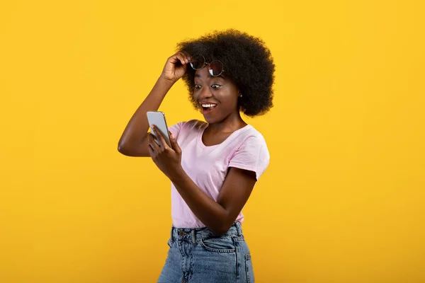 Surprised african american woman looking at smartphone and lifting her sunglasses over yellow studio background — Stock Photo, Image