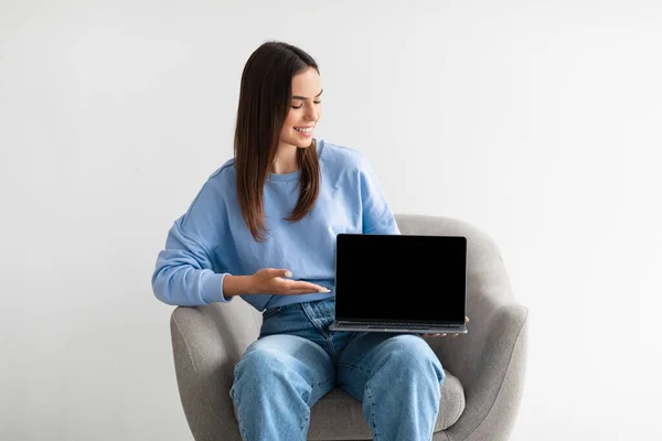 Leende kvinna visar laptop med tom skärm, sitter i fåtölj på vit studio bakgrund, mockup — Stockfoto