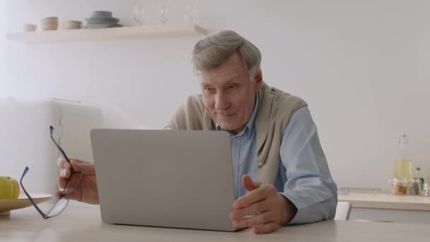 Pozytywny senior patrzący na laptopa, siedzący we wnętrzu kuchni, oglądający wiadomości online, korzystający z nowoczesnych technologii — Wideo stockowe