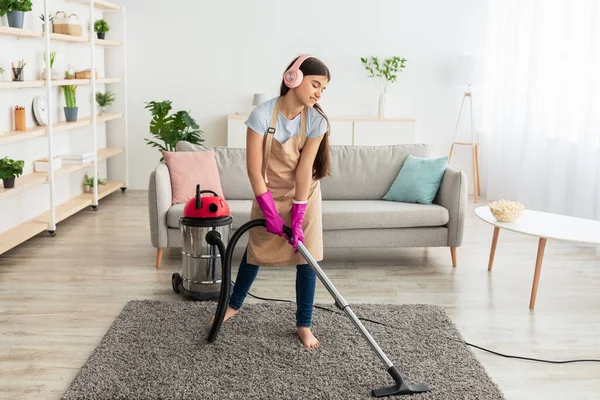 Happy Indian teen girl cleaning her home, wearing headphones, listening to music while vacuum uming floor, having fun — стоковое фото