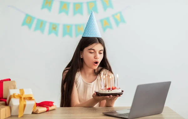 Indiska tonåring flicka i festlig hatt har online födelsedagsfest, blåser ljus på tårta framför laptop webbkamera — Stockfoto